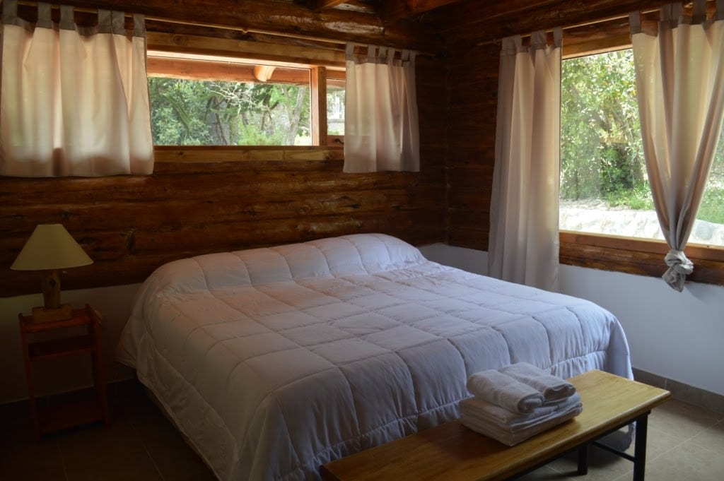 Dormitorio de planta baja de Cabaña 1 con cama matrimonial, dos ventanas y banco de madera al pie.